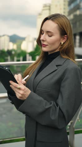 a businesswoman in a gray suit inspects her surroundings, then writes notes on a digital tablet with a stylus outside modern office buildings