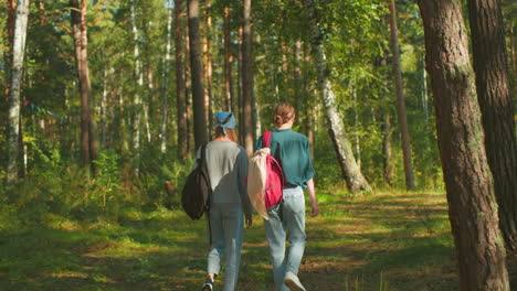 back view of two sisters walking through lush green forest, one wearing blue hair tie and carrying backpack, other with cloth draped over bag and hair tied back