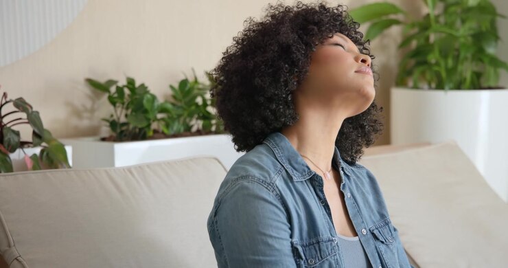 Donna afroamericana con camicia di jeans su divano chiaro, che chiude gli occhi e ruota il collo per rilassarsi vicino a una fioriera.