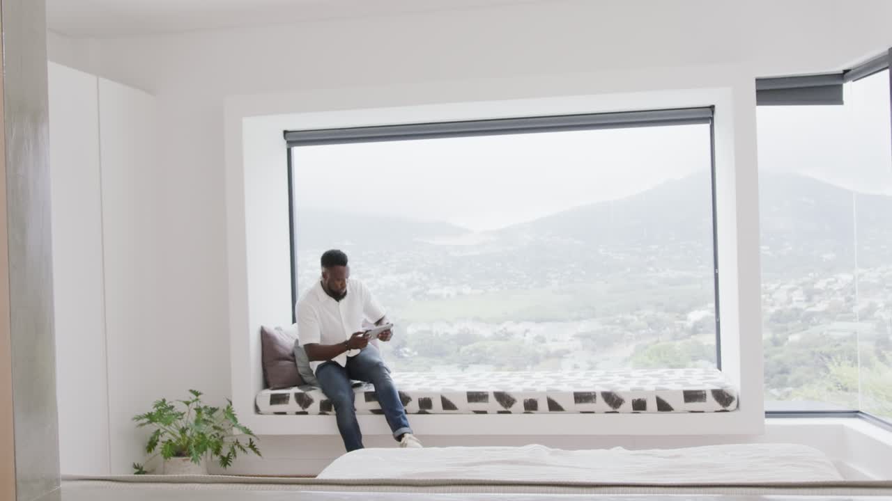 African man holding phone, moving to sit on window bench, reclining on pillows viewing phone