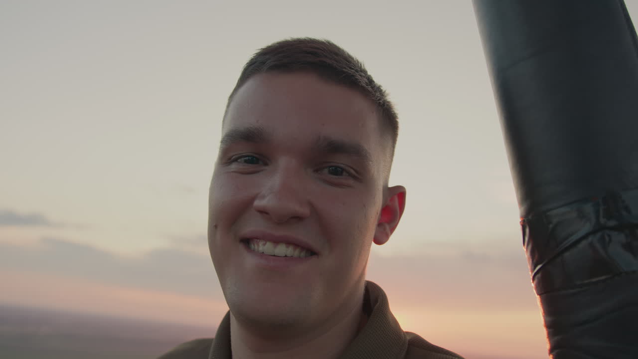 young man gazes upward and smiles at camera from hot air balloon basket under pastel sunset sky over expansive fields while gentle evening breeze creates intimate airborne portrait moment