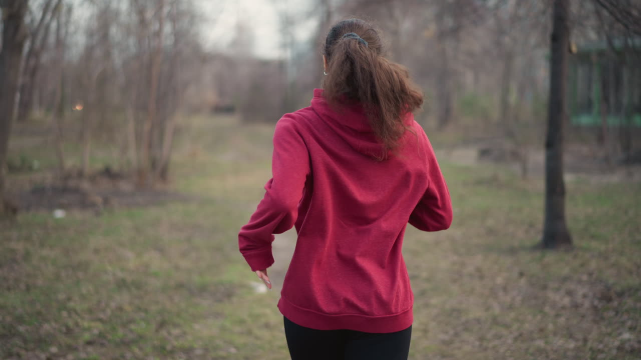 White Female In Hoodie Enjoying Evening Run, Single Woman In Red Hoodie Jogging Across Open Park At Dusk, Solitary Woman Runs Through Leafless Park Wearing Red Hoodie During Twilight Hours