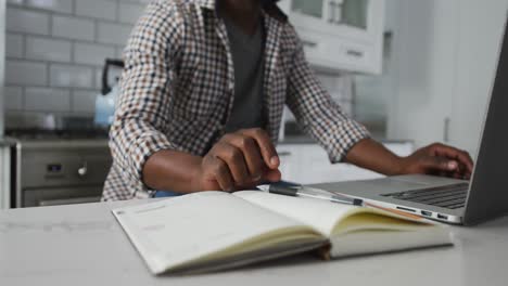 midsection of african american man working at home in kitchen writing and using laptop