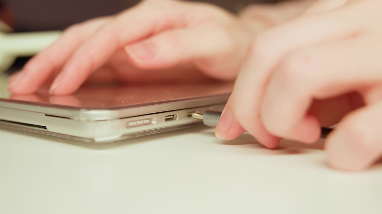 Close-up of hands interacting with slim tablet or laptop at desk edge showing tapping, swiping, and cable connection; fingertips display motion blur from active touch while connector is inserted