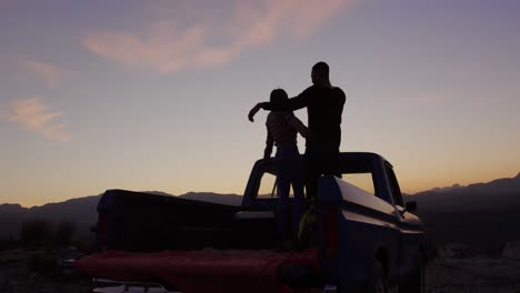 Young-couple-on-a-road-trip-sitting-outside-on-their-truck-at-dusk