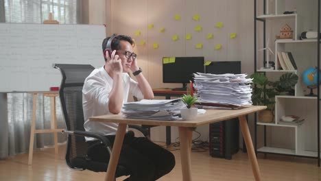 asian man wearing headphones and listening to music on smartphone after working with documents at the office