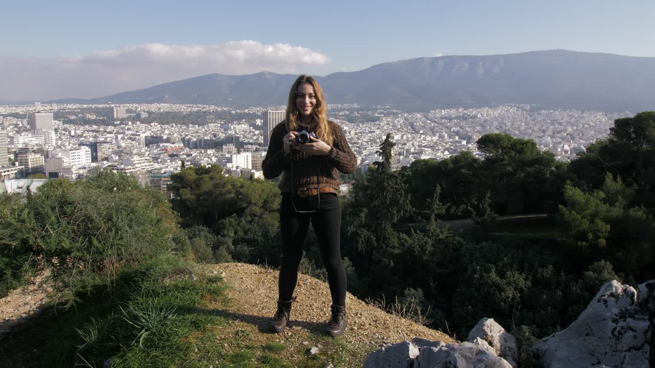 una mujer joven de pie en la colina de lycabettus toma una foto con su cámara analógica con vista de atenas, grecia en el fondo, pov gimbal