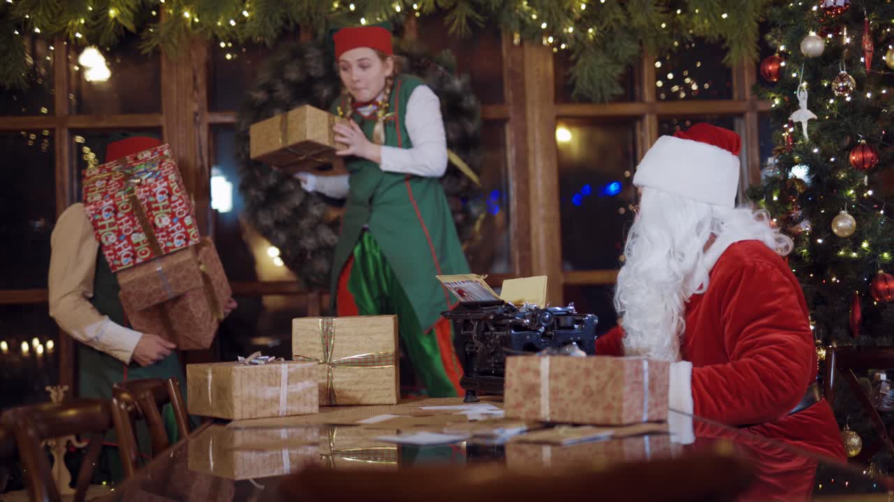 Santa quarrels on naughty elves inside the Christmas decorated room. Two elves having fun with presents in boxes but Santa Claus teaches them.