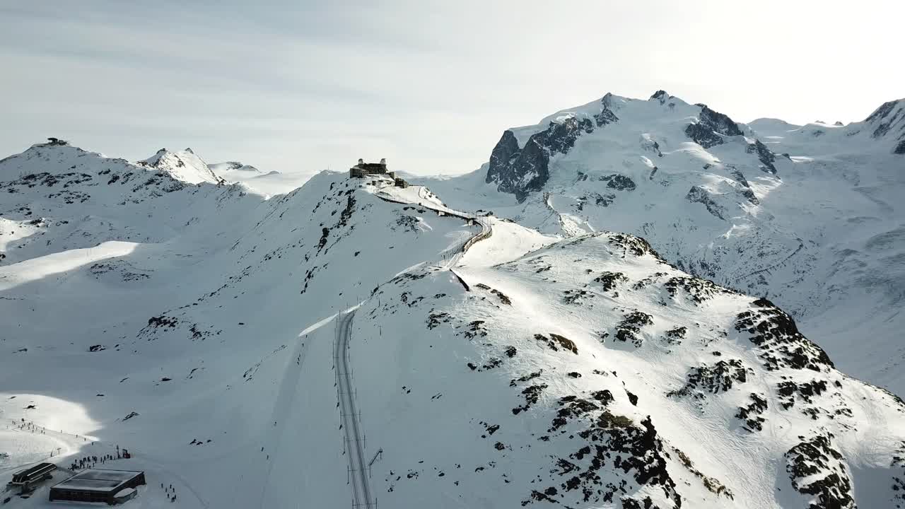 Drone view of Gornergrat, flying straight ahead over snow-covered mountains, revealing rugged peaks and a lone building perched on a ridge amidst the vast, icy Swiss Alpine landscape