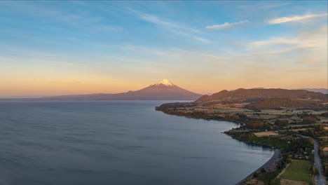 Eine-Ruhige-Luftaufnahme-Zeigt-Einen-Riesigen-See-Mit-Einem-Schneebedeckten-Osorno-Vulkan-In-Der-Ferne-Bei-Sonnenuntergang