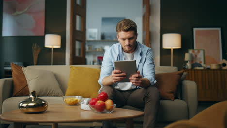 calm man browsing tab device at home. serious guy sitting sofa scrolling tablet