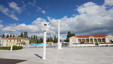 a spacious plaza with white columns, surrounded by buildings with red-tiled roofs, trees, and a clear blue sky