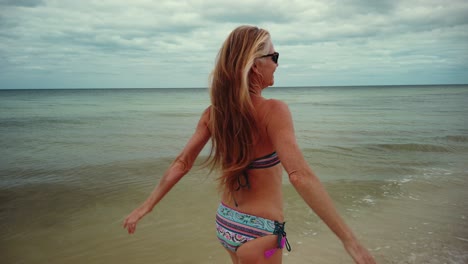 riendo, mujer madura juguetona en bikini y gafas de sol se da la vuelta y corre en la playa