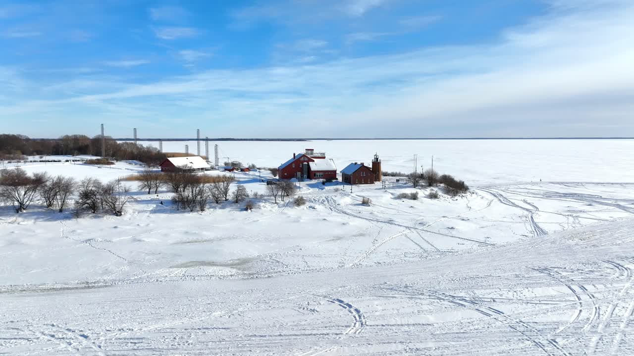 Una ripresa aerea a bassa quota che mostra tradizionali edifici in legno rosso e un faro a Capo Ventė, circondato da una laguna ghiacciata e neve alta.