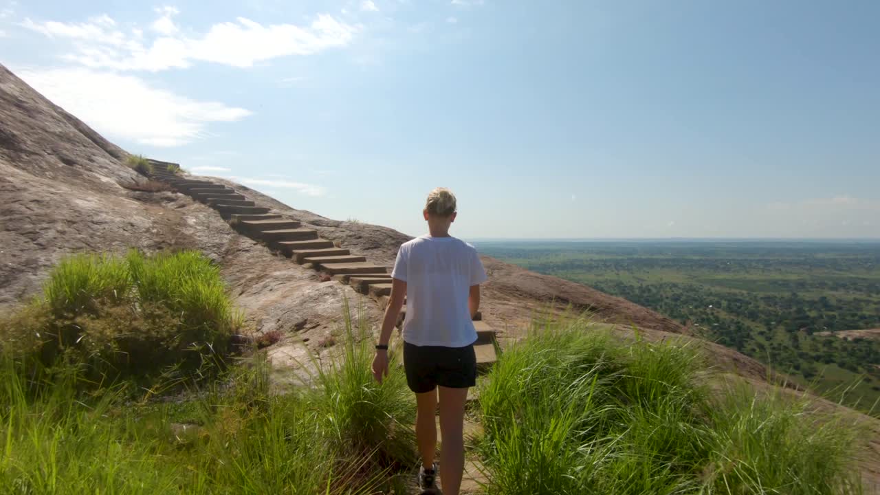 una foto de seguimiento de una mujer caucásica rubia subiendo escaleras de hormigón en la cima de una gran montaña africana de granito con vistas increíbles