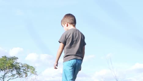 Caucasian-boy-toddler-watching-the-ground-and-stands-up-walking-,-low-ground-level-view,-with-beautiful-blue-sky-in-the-background-120fps