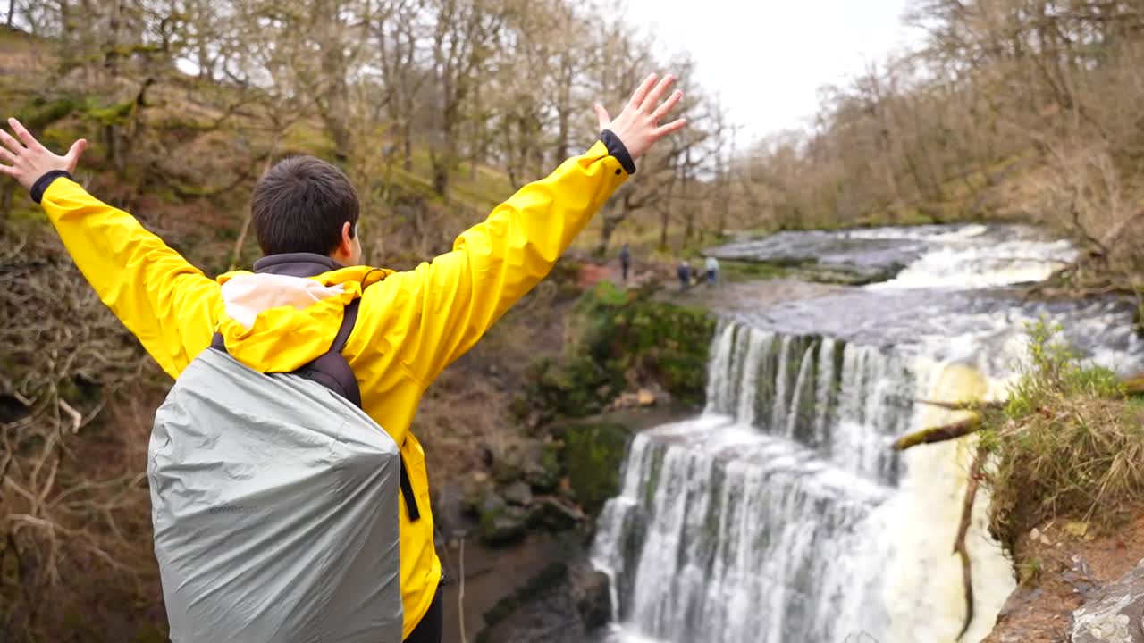 hombre levantando los brazos con una impermeable amarilla y una bolsa cubierta frente a una gran cascada en sgwd isaf clun-gwyn, gales