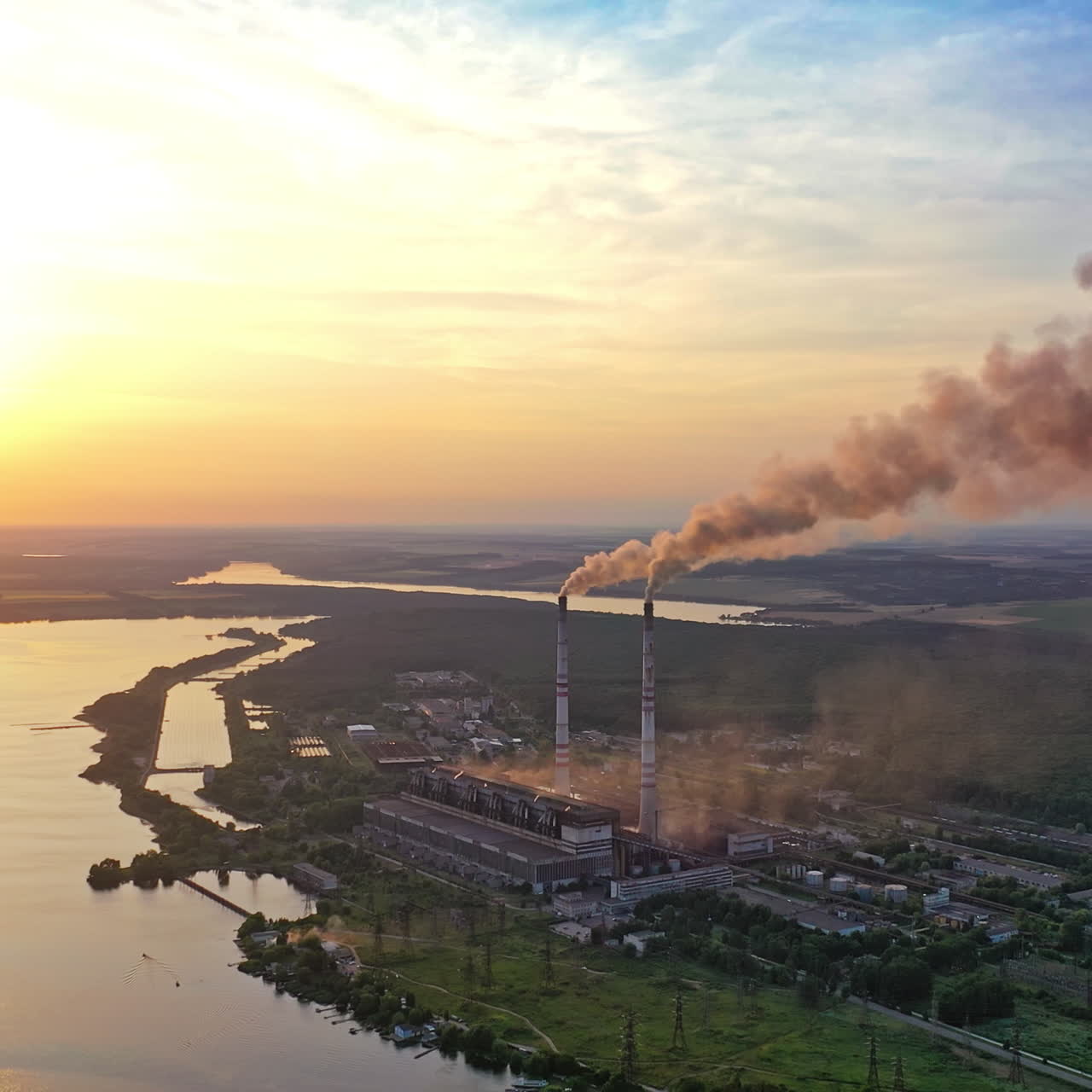 Smoke from pipes on factory near the river at sunset. Air pollution from industrial plant on the natural background in the evening.