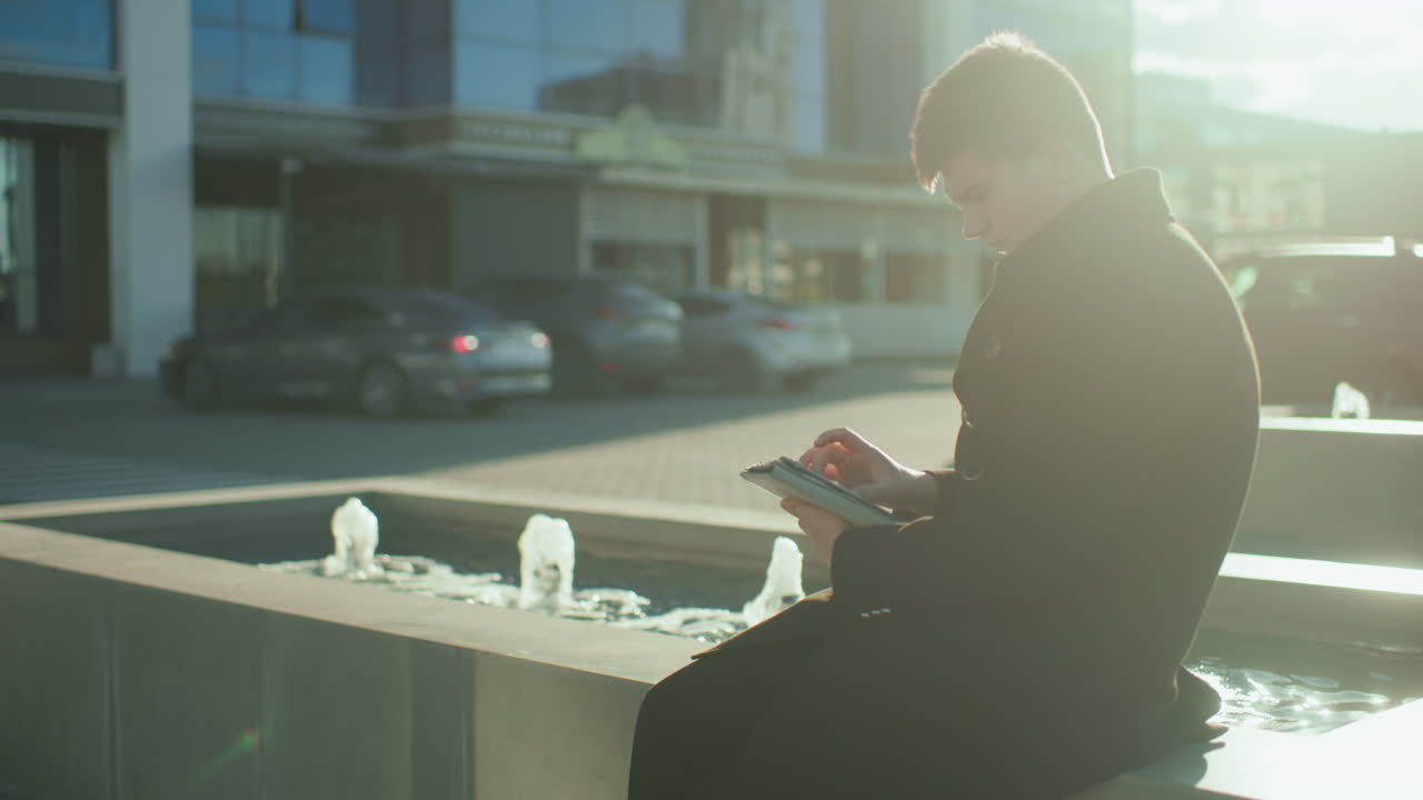 Man operates tablet near flowing spring water fountain while a car drives away in blurred background, illuminated by soft sunlight and urban building