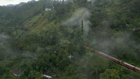 Establishing-Aerial-Drone-Shot-of-Red-Train-Through-the-Trees-in-the-Hills-in-Ella-Sri-Lanka-on-Misty-Morning