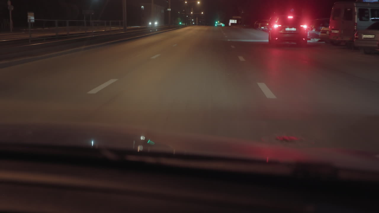 Car driving through quiet street at night with headlights and tail lights from multiple vehicles visible, deserted road under glow of street lamps
