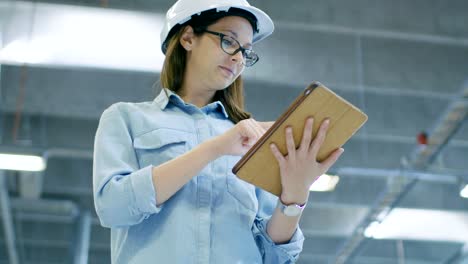 female industrial engineer in a hard hat uses tablet computer while walking in big factory.