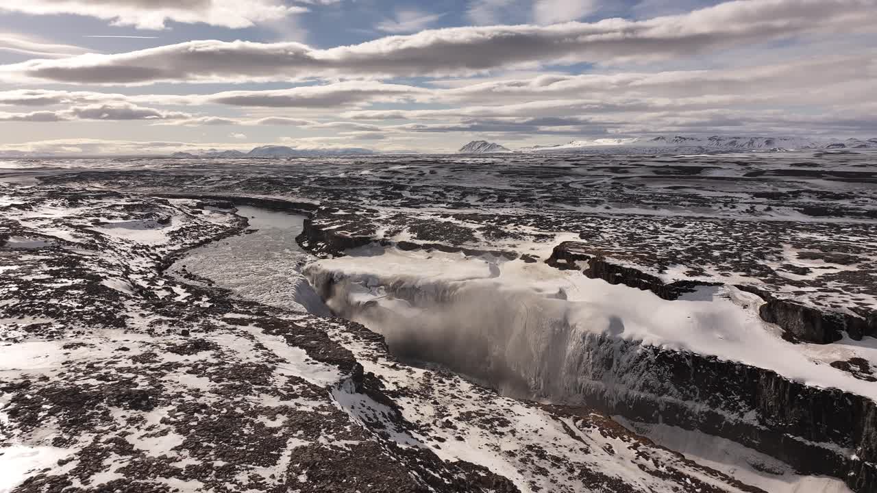 Aerial view of a powerful waterfall in a snowy, rugged landscape, likely Dettifoss in Iceland. Icy river flows into a deep gorge.