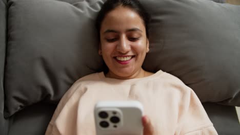 Close-up-a-happy-brunette-girl-in-a-light-jacket-leans-her-elbows-and-lies-down-on-a-gray-sofa-on-a-pillow-while-relaxing-and-communicating-on-social-networks-using-a-white-smartphone-at-home