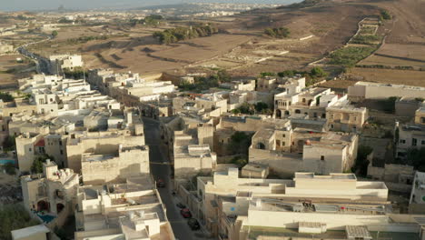 Road-through-small-Town-on-Gozo,-Malta-Island-with-Car-traffic-in-between-Beige-and-Brown-Colored-Houses,-Aerial-View-from-Above,-Dolly-forward