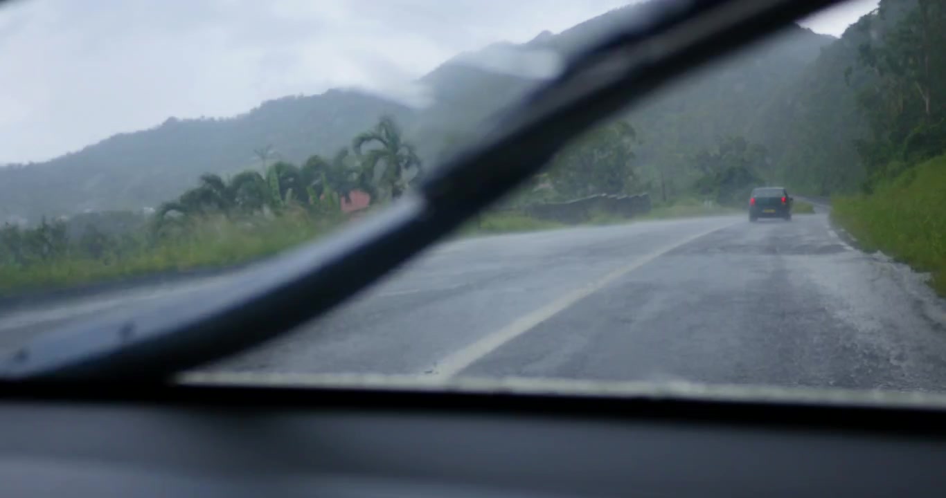 Driving car in heavy summer rain. View through wet windscreen with wipers on. On the road in a Caribbean countryside.
