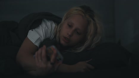 una chica rubia con una camiseta blanca de repente se despierta mira ansiosamente al teléfono y se levanta de la cama temprano en la mañana