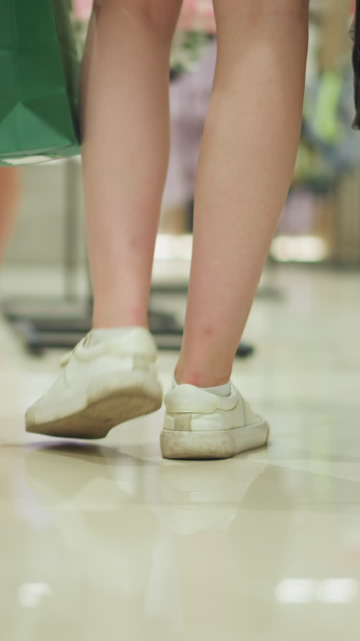 캔버스 신발을 입고, 다채로운 쇼핑 가방을 들고, 잘 조명된 쇼핑몰에서 끔하게 배열된 옷을 고, 배경에 다른 쇼핑객과 함께 고 있는 여자의 뒤쪽 다리 시각