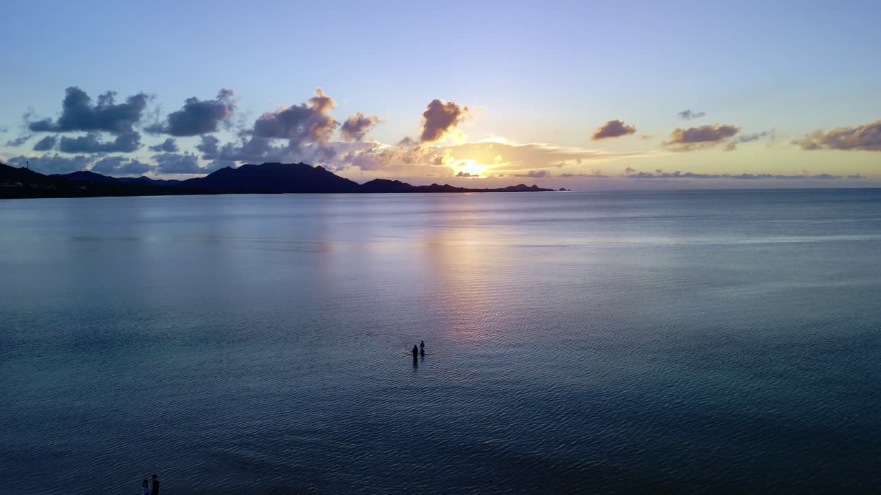 Cinematic shot of figures on a beach and in the water at dusk, with the setting sun reflecting on a mirror-like ocean surface and mountain silhouettes