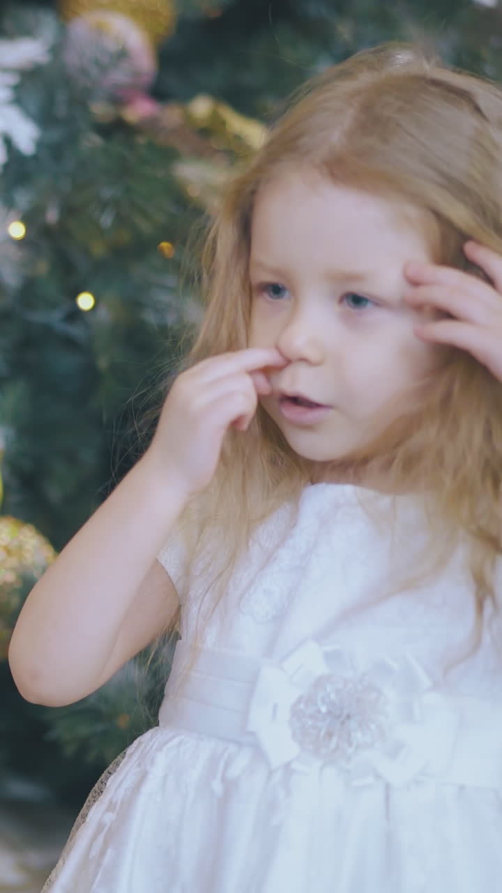 funny little blond girl in elegant dress touches balls on beautiful decorated Christmas tree at home