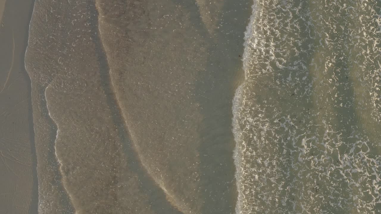 Aerial ascending slowly on rolling waves breaking on sandy sunlit beach