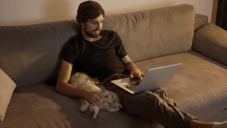 Hombre-Feliz-Trabajando-En-Una-Computadora-Portátil-En-Casa-Mientras-Se-Sienta-En-El-Sofá-Y-Acaricia-A-Un-Gato