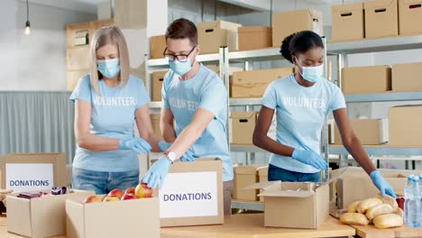 Grupo-Multiétnico-De-Voluntarios-En-Cajas-De-Embalaje-De-Máscaras-Faciales-Con-Comida-Y-Agua-En-Un-Almacén-De-Caridad