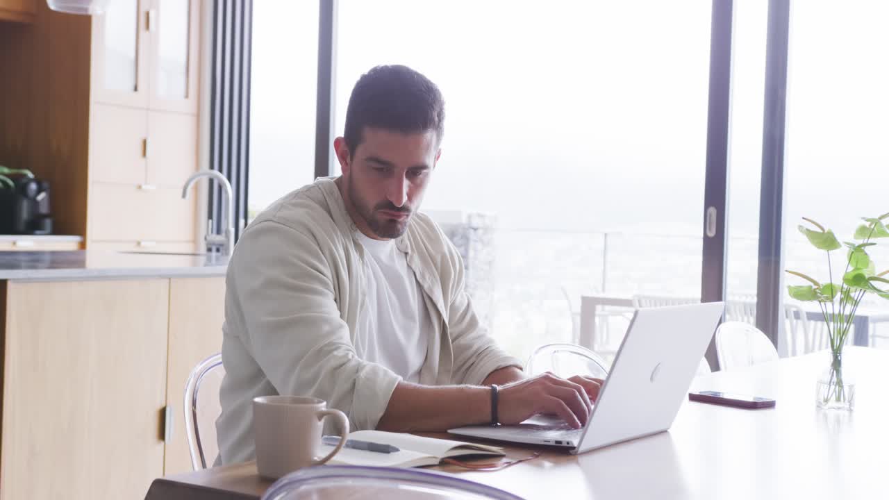 Hombre adulto teletrabajando en la mesa de la cocina junto a puertas de cristal, escribiendo en un portátil tras revisar unos papeles.