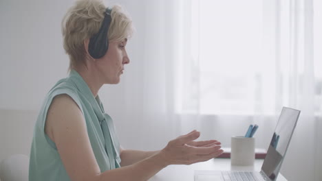 una mujer mayor se está comunicando con colegas o clientes a través de un chat en línea en una computadora portátil hablando y escuchando con auriculares