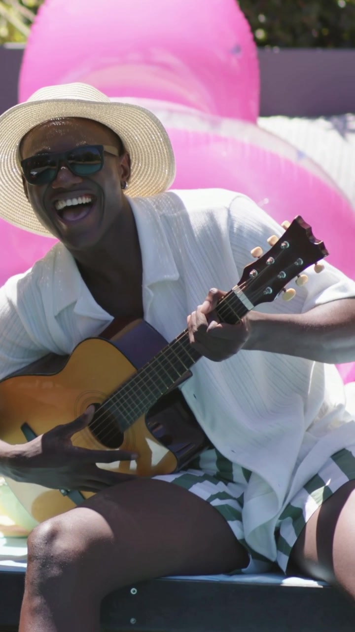 Video verticale: Uomo con cappello di paglia e occhiali da sole che strimpella la chitarra vicino a un galleggiante rosa, mentre la telecamera si alza per divertimento.