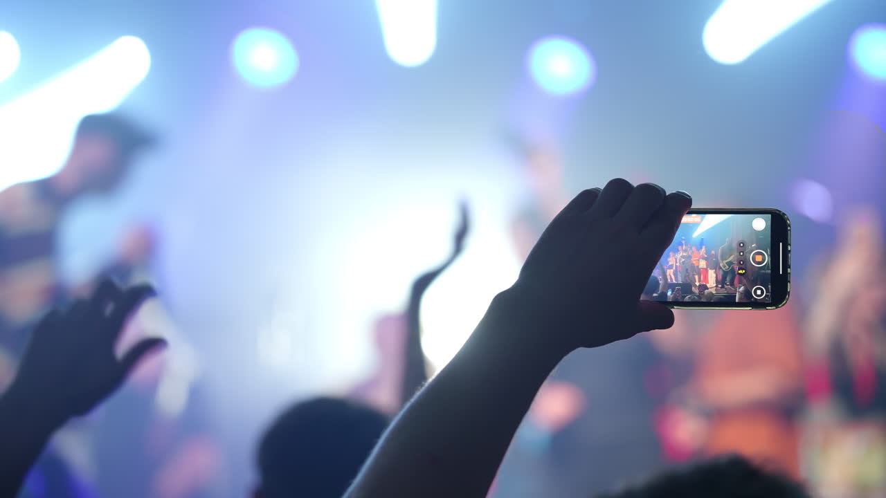 Fan recording live music concert with smartphone, standing silhouetted against vibrant stage lights, capturing dynamic performance moment with digital technology and personal passion