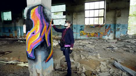 young man professional graffiti painter is working inside abandoned building, he is painting with spray aerosol paint on high column. modern art and creativity concept.