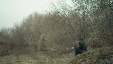 a close view of a man in a green jacket sitting alone on a grassy hillside, surrounded by bare trees, operating a phone