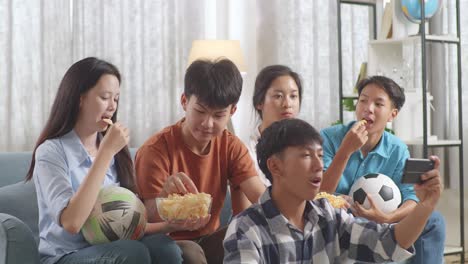 asian teenagers cheering, eating and watching football game on smart phone at home