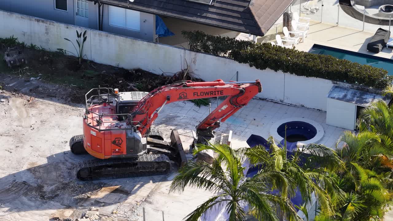 An orange excavator collects and loads demolition debris into a blue dumpster on a cleared residential lot in bright daylight, viewed from above