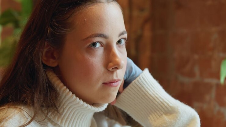 Close up of young woman with long brown hair wearing white turtleneck sweater resting her head on one hand in indoor setting. background features warm lighting and blurred brick walls