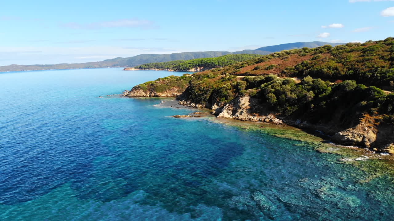 Aerial drone view of the Aegean sea rocky coast with greenery along the shore and hills in the distance, blue water in Greece