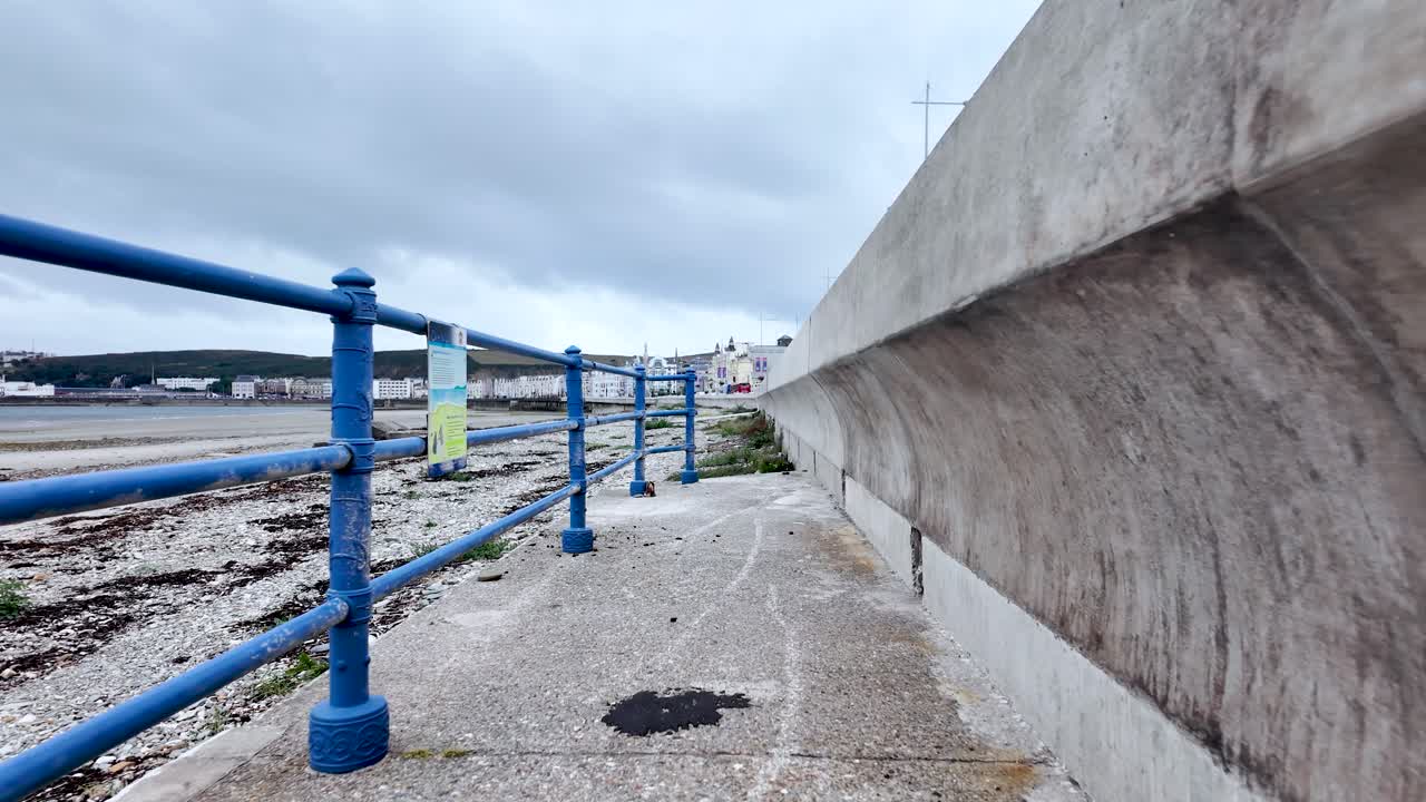 Overcast coastal scene of a concrete path, blue railings, and a sea wall along Douglas Beach