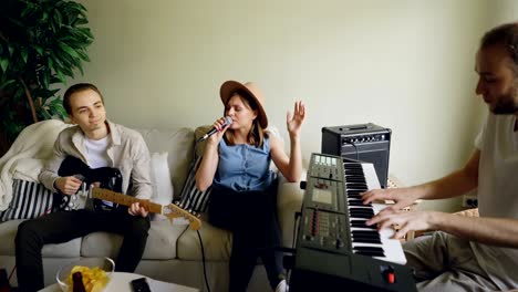 cheerful friends musicians are practising in the house, female vocalist is singing in microphone and men are playing the keyboard and the guitar together.