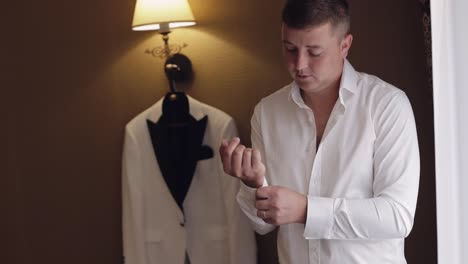 apuesto novio vestido en la mañana de la boda y arregla sus botones en una manga de camisa blanca, en el interior